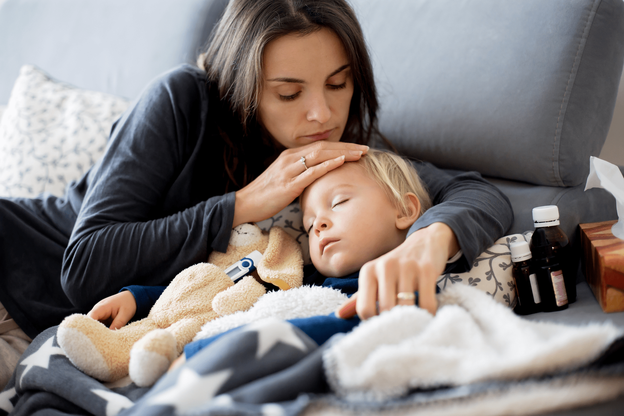 woman holding a tired-looking child, surrounded by blankets and a teddy bear